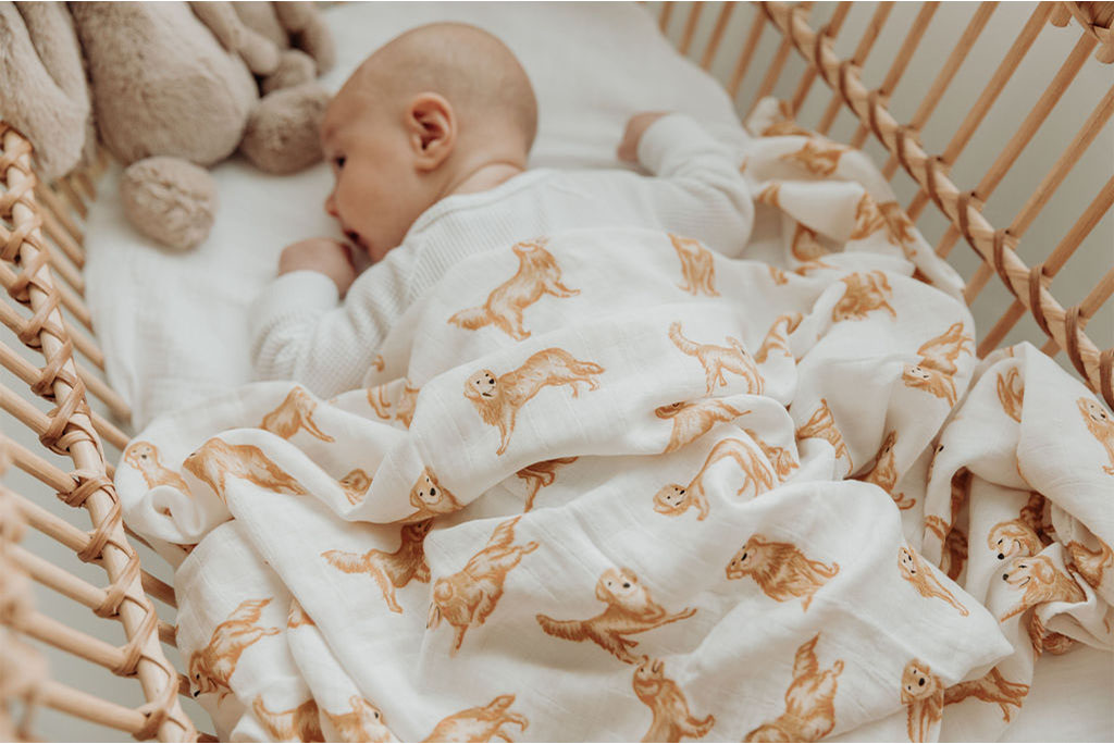 Baby in bassinet covered with swaddle printed with a golden retriver. Jelly cat bunny in cot